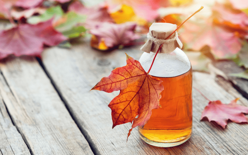 A small jar of maple syrup stands on a wooden surface, framed by vibrant fall leaves in the background; a single orange maple leaf rests delicately on the jar's lid.