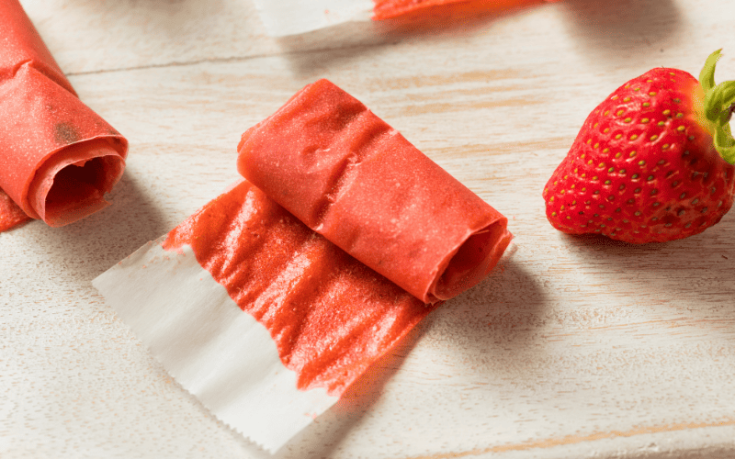 Delicious fruit leather is artfully presented on parchment paper atop a wooden countertop, accompanied by a fresh, vibrant strawberry placed tastefully to the side.