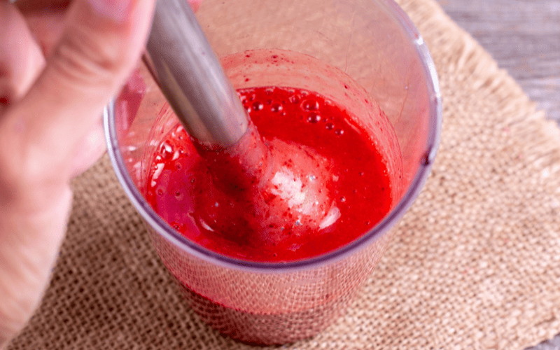 Top-down view of a person using a stick blender to puree fresh strawberries into a vibrant and smooth mixture.