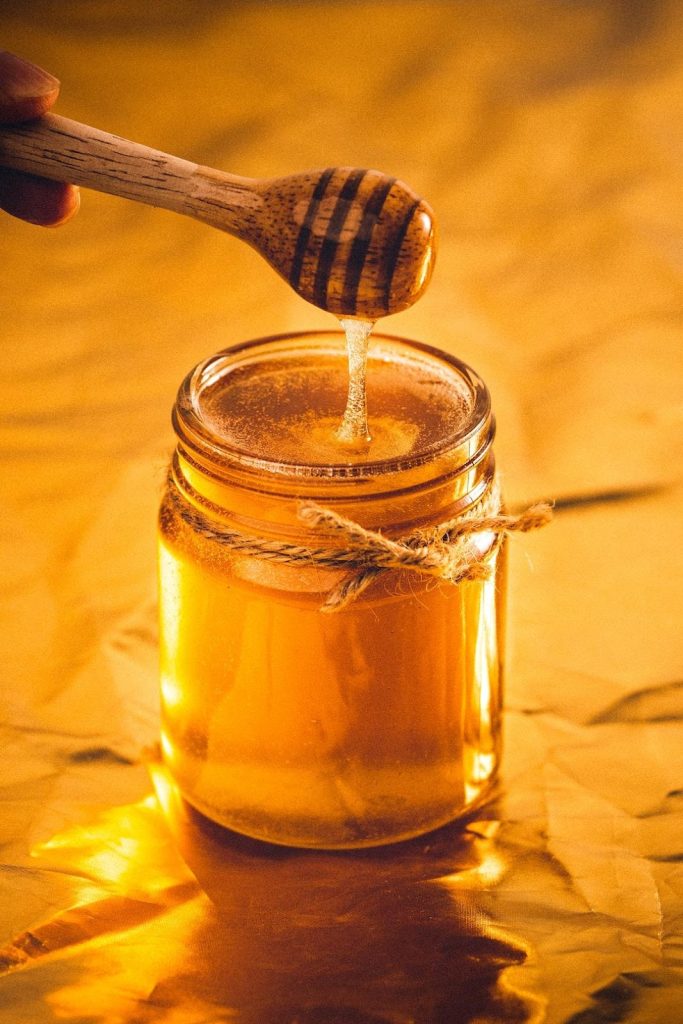 A glass jar of honey with a metal lid is placed on a flat surface. A wooden spoon is suspended a few inches above the jar, and a steady stream of golden honey flows from the spoon, creating a glistening arc as it descends into the jar. The backdrop features a soft, muted texture. The overall scene captures the process of honey being drizzled into the jar, showcasing its rich color and sticky fluidity.