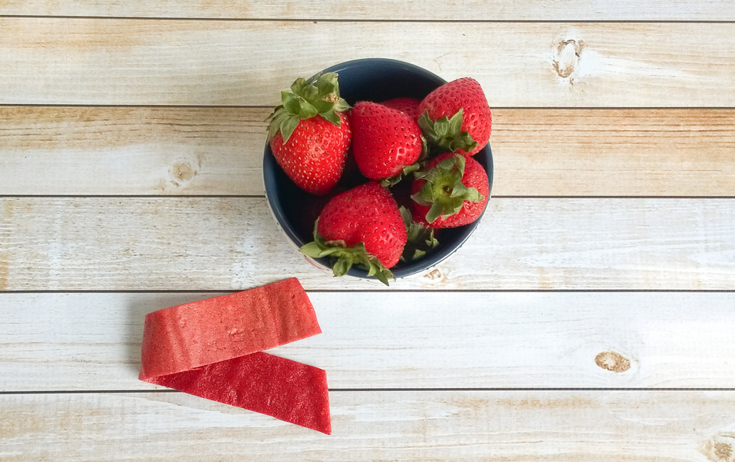 Overhead view of a luscious strawberry fruit roll-up placed beside a bowl of fresh strawberries on a rustic wooden countertop.